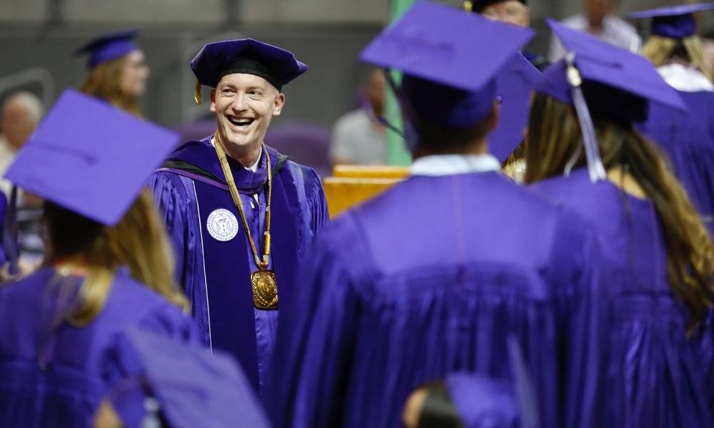 校长维克多J. 小Boschini.她面带微笑，身着校服，向戴着紫色帽子、身穿紫色长袍的TCU毕业生致意